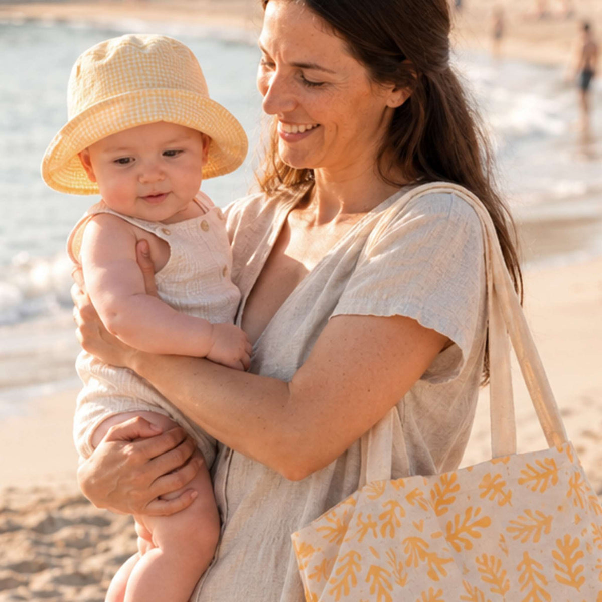 Une maman et son bébé à la plage, avec un tote bag Biolane et un bob